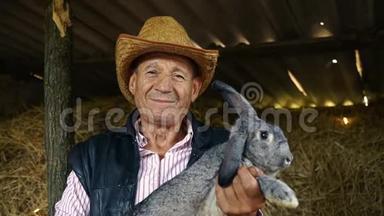 一个戴草帽的<strong>老农</strong>夫正抱着一只灰色的大兔子。 干草背景下男人的肖像